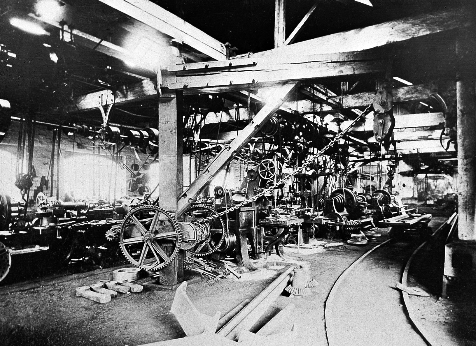 A circa-1890 machine shop with overhead shafts and belts descending to machines, surrounded by pulleys and large gears.